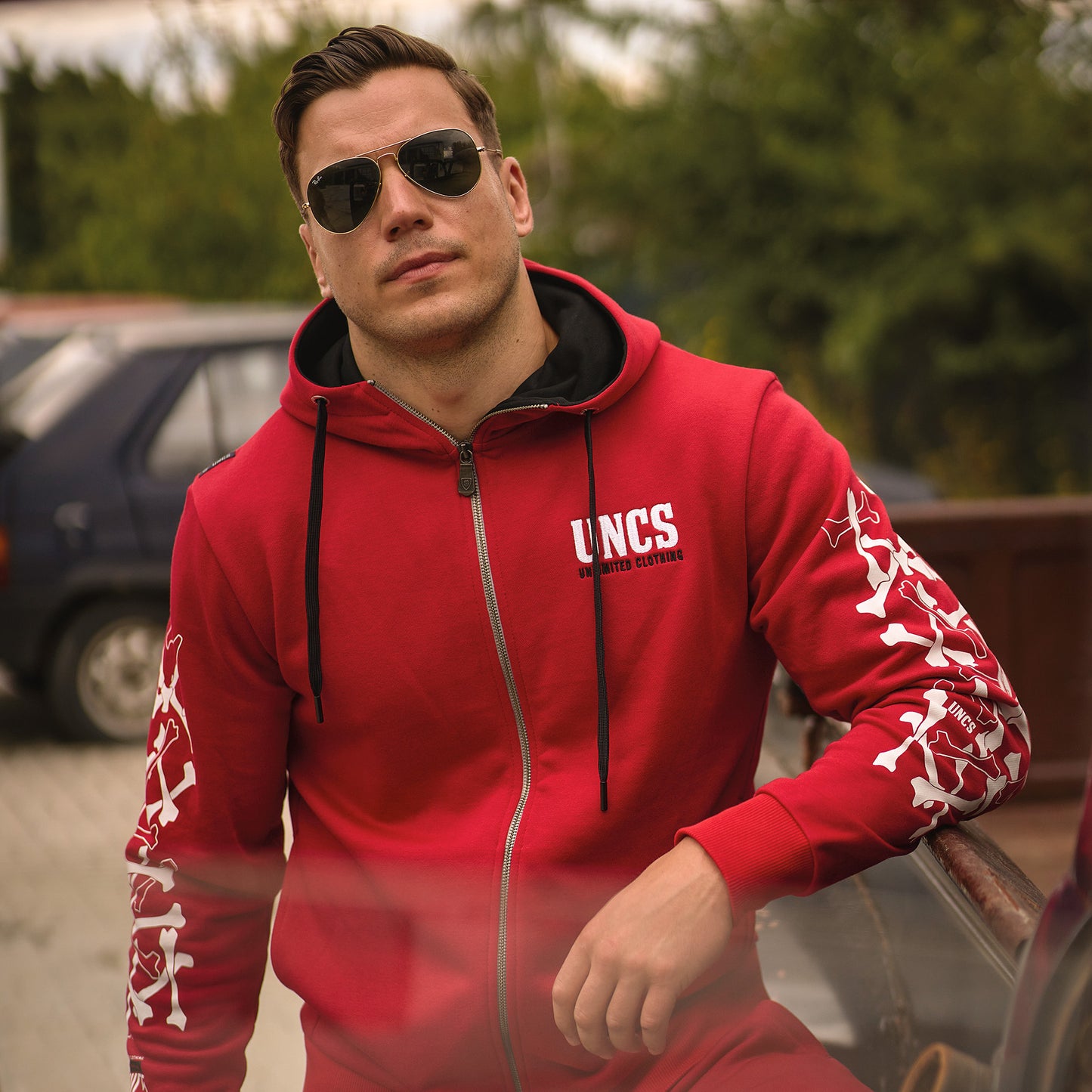 Muž v červené mikině UNCS s bílými prvky s autem a stromy v pozadí.