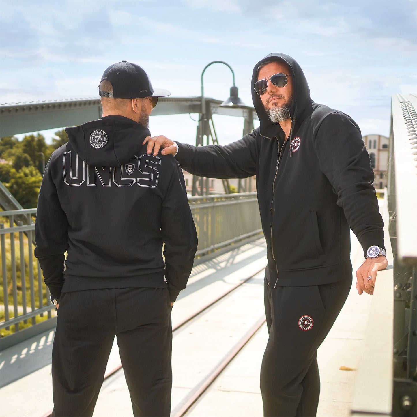 Dva muži v černých teplákových setech UNCS na mostě s parkem a budovou v pozadí.