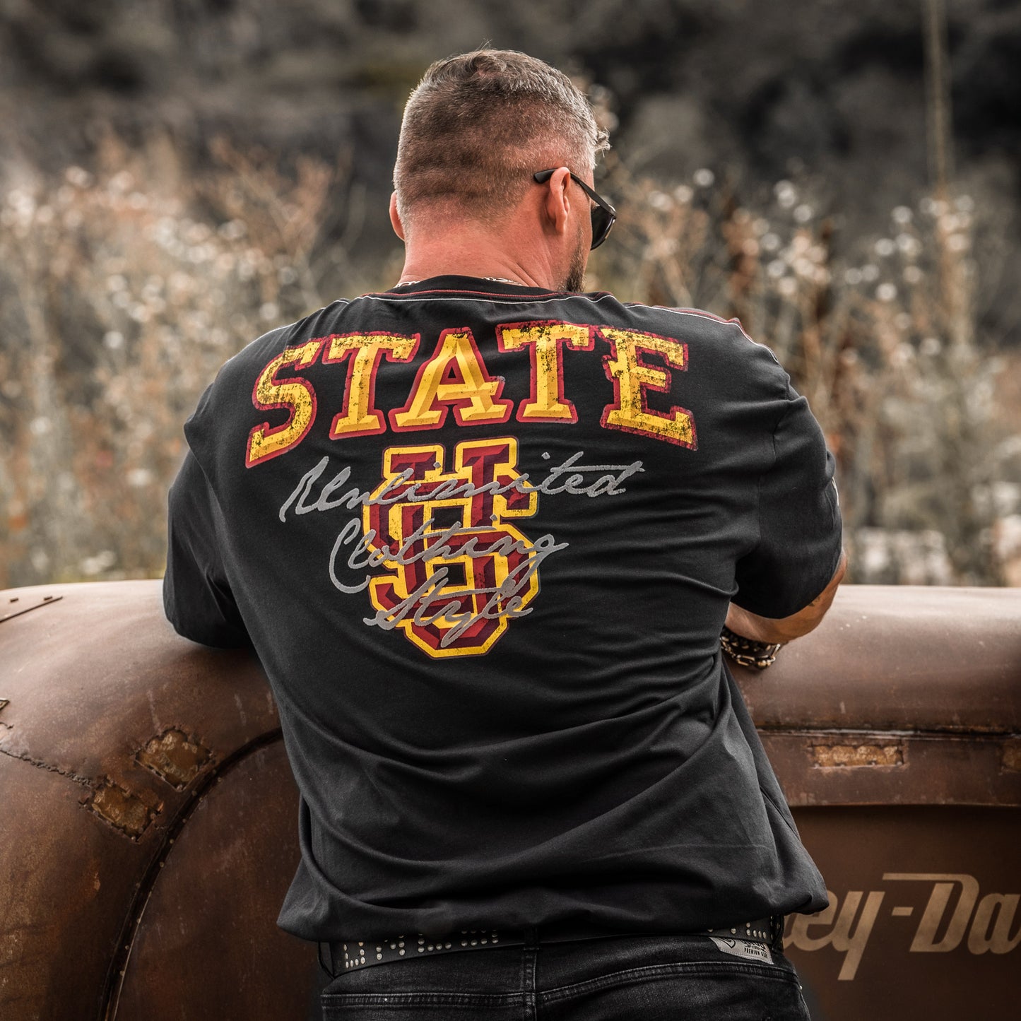 Muž v černém tričku UNCS State s vintage logem na zádech, motorkářský styl, opřený o staré auto v přírodě.